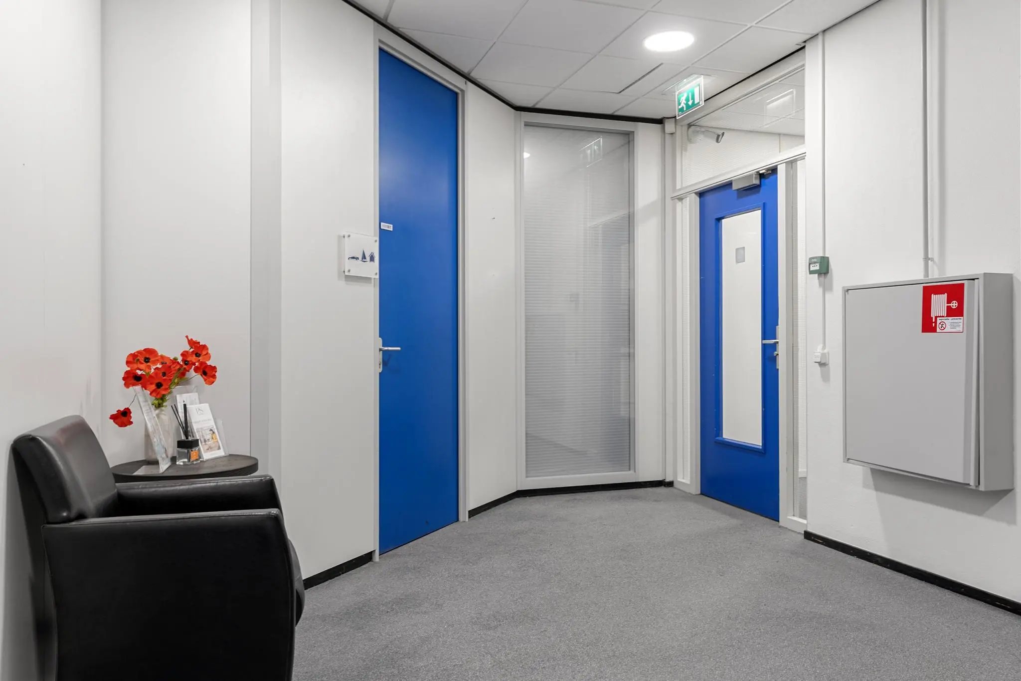 Witte gang met grijze vloerbedekking, felblauwe deuren, een zwart stoeltje en een tafeltje met bloemen en folders in een kantoor- of praktijkruimte aan de Huis-ter-Heideweg.